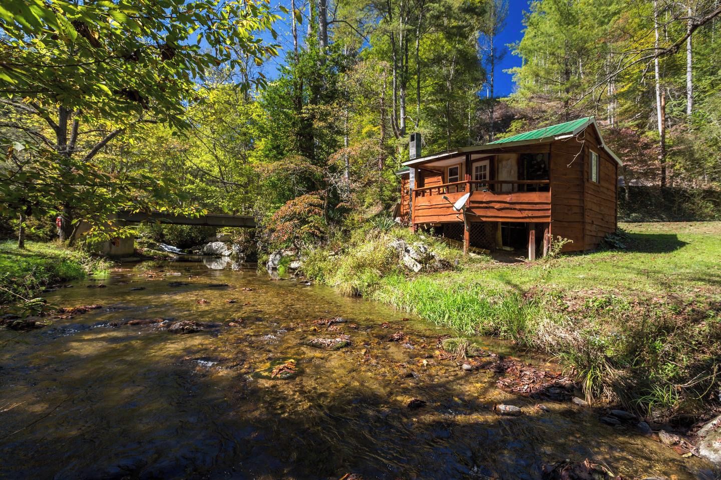 A wooden cabin with a green roof, situated by a clear stream surrounded by trees.
