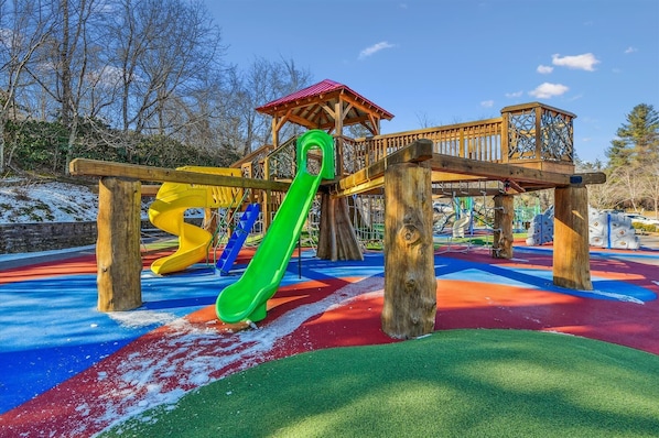 Un parque infantil con una estructura de madera para jugar, que incluye un tobogán y elementos para escalar, sobre una superficie de caucho de colores.
