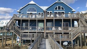 Une maison de deux étages située en bord de mer, dotée d’une terrasse en bois et de plusieurs balcons.