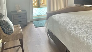 Une chambre à coucher comprenant un lit, un téléviseur, un ventilateur de plafond, une fenêtre avec des stores, une petite table sur laquelle est posée une statue, et un fauteuil en osier.