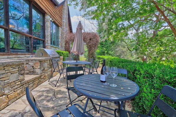 An outdoor patio with stone walls, a grill, and a table with chairs and a wine glass.