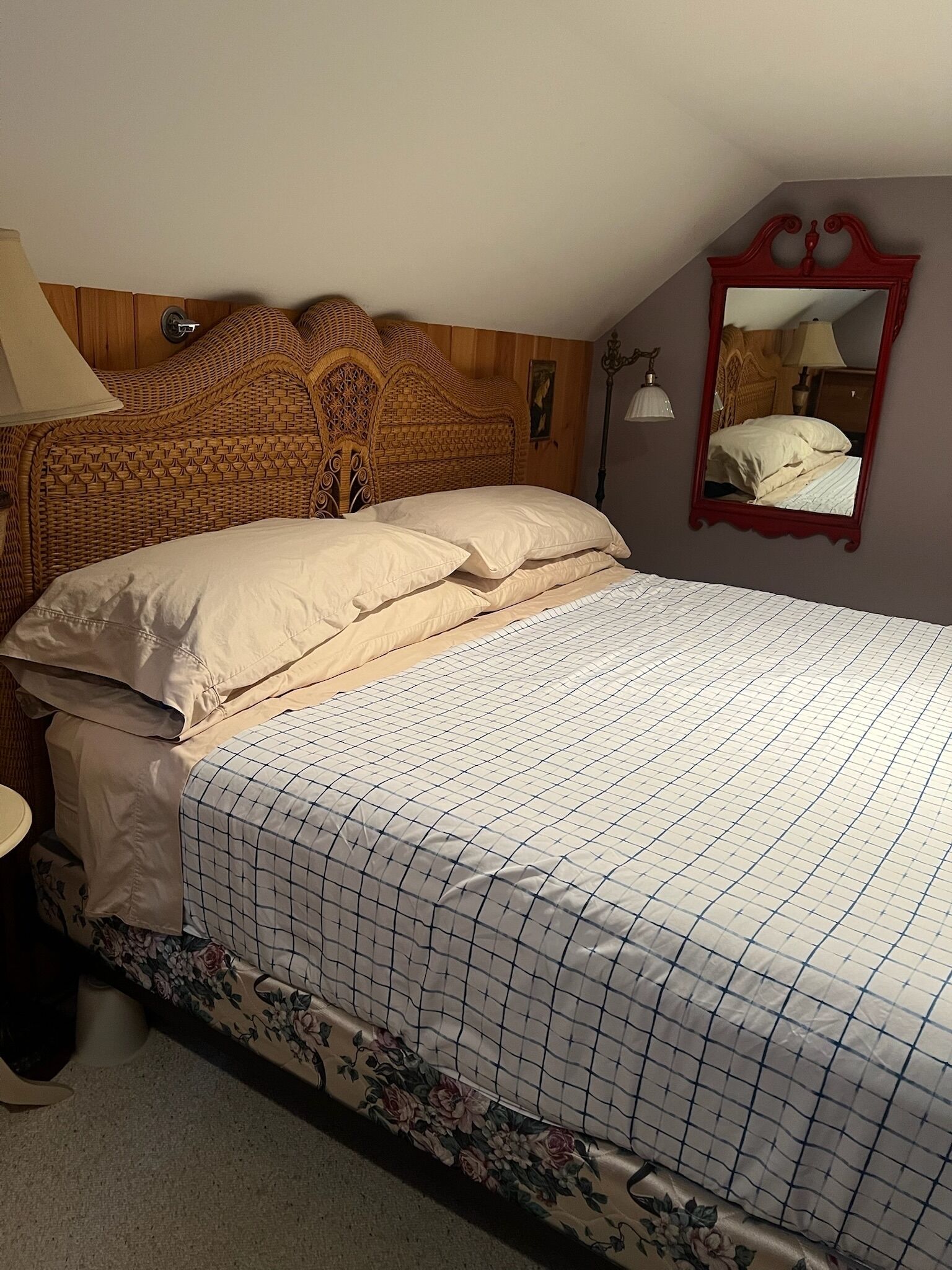 Una cama bien hecha con una colcha estampada y falda floral, un cabecero de madera y un espejo en la pared.