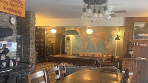 Zona pranzo con un tavolo di legno, sedie, una mappa appesa al muro e un angolo bar con un orologio e un cartello.