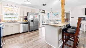 A modern kitchen with stainless steel appliances, a central island, and a dining area with a high chair. - Minutes to Clearwater Beach, Indian Rocks Beach- Sleeps 10 w/Firepit & BBQ! (Largo)