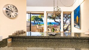 A kitchen with granite countertops, a clock, and a view of the ocean through large windows. - Oceanview Family Favorite! Beach, Pool, Hot Tub, AC + Video Tour (Kihei)