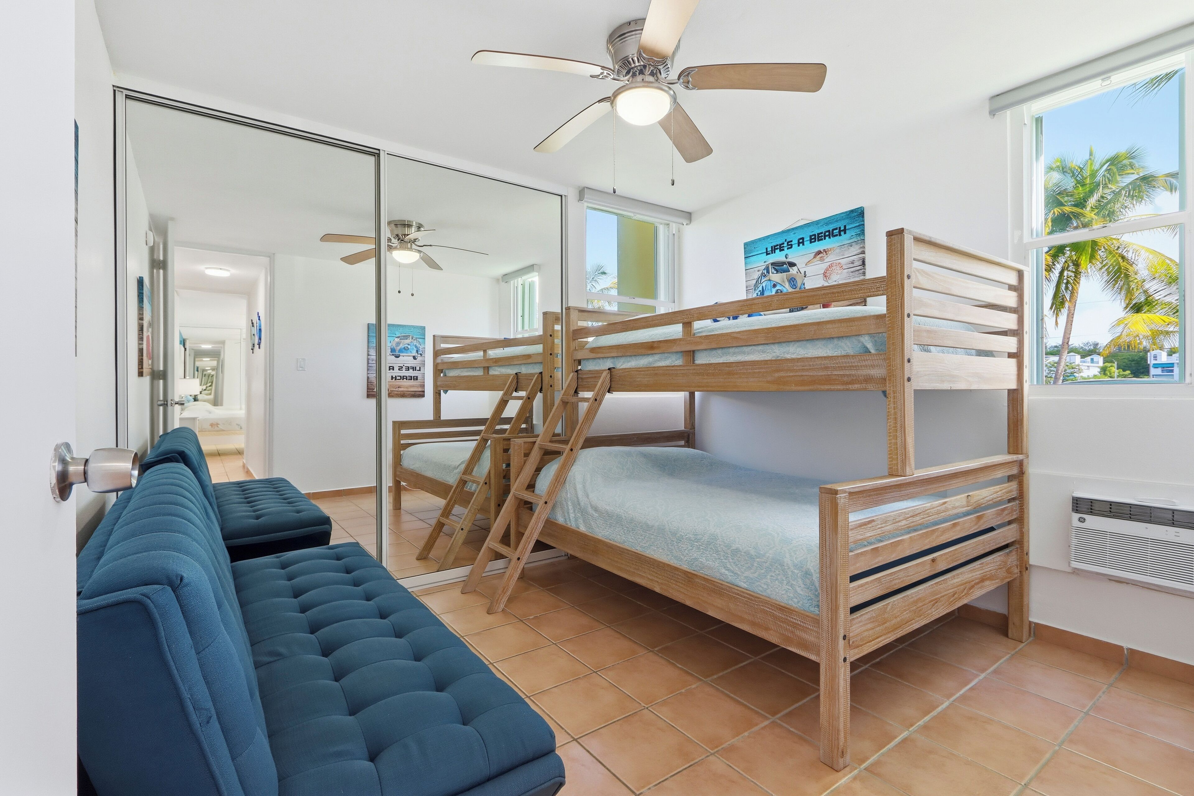 Una cama litera con escalera, un sofá azul, un ventilador de techo y un cartel en la pared.