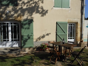 A rustic outdoor seating area with a wooden table and chairs, a potted plant, and a door with green shutters.