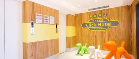 A hotel reception area with a wooden wall, a reception desk, and a sign for the Click Hotel. There are three small, colorful animal-shaped decorations on the floor.