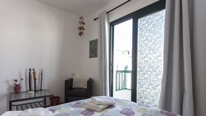 Una camera da letto con un letto, una sedia, un tavolino con oggetti decorativi e vista su un balcone attraverso una finestra.