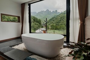 Un baño moderno con una bañera independiente, vistas a un paisaje montañoso y un ventanal con cortinas.