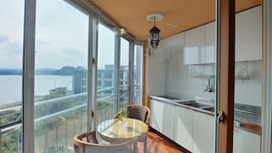 Een balkon met een glazen tafel en stoelen, uitzicht op een wateroppervlak, en een keuken met witte kasten en een fornuis.