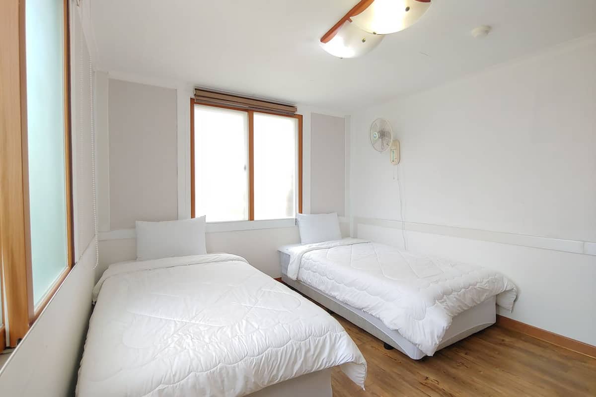 A room with two single beds, a window with blinds, a wall-mounted fan, and a ceiling light fixture.