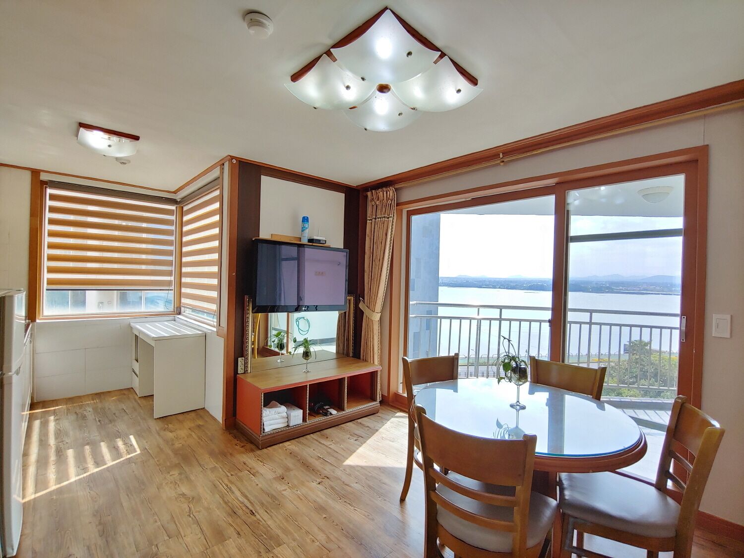 A room with a balcony, a TV, a dining table, and a view of a body of water.
