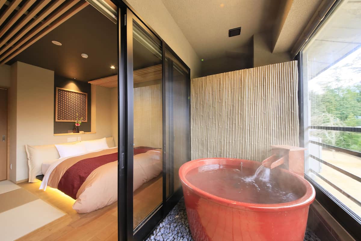 A modern hotel room with a large sliding glass door leading to a balcony, a red hot tub, and a bed with a red blanket.