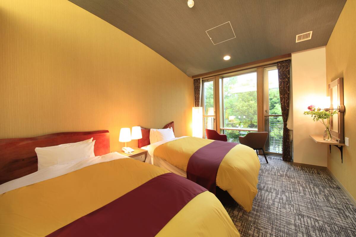 A hotel room with two beds, a nightstand with a lamp, a chair, a window with curtains, and a small table with a vase.