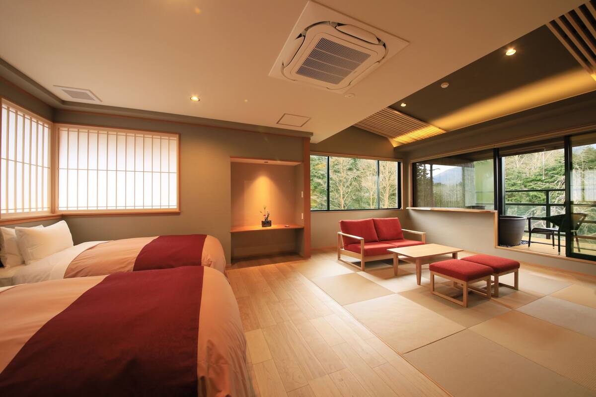 A hotel room with two beds, a sofa, a table, and large windows offering a view of greenery.