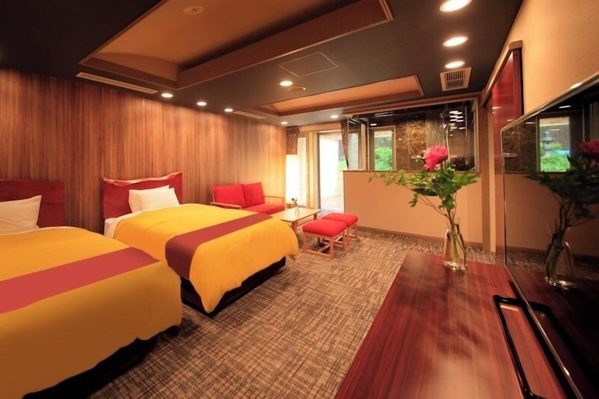 A hotel room with two beds, a small table, a chair, and a large window.