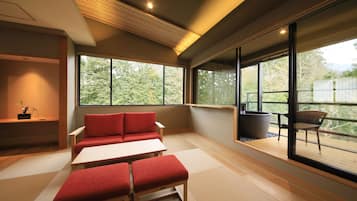Un interior moderno con un sofá, otomanas y una mesa. Amplias ventanas ofrecen vistas a árboles y un balcón.