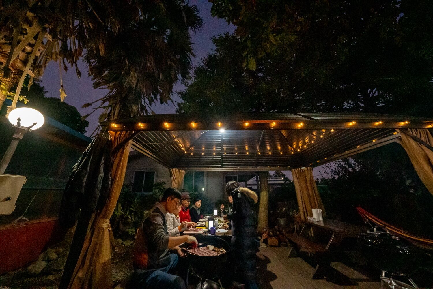 An outdoor dining area with a covered seating arrangement, string lights, and a view of the surrounding trees.