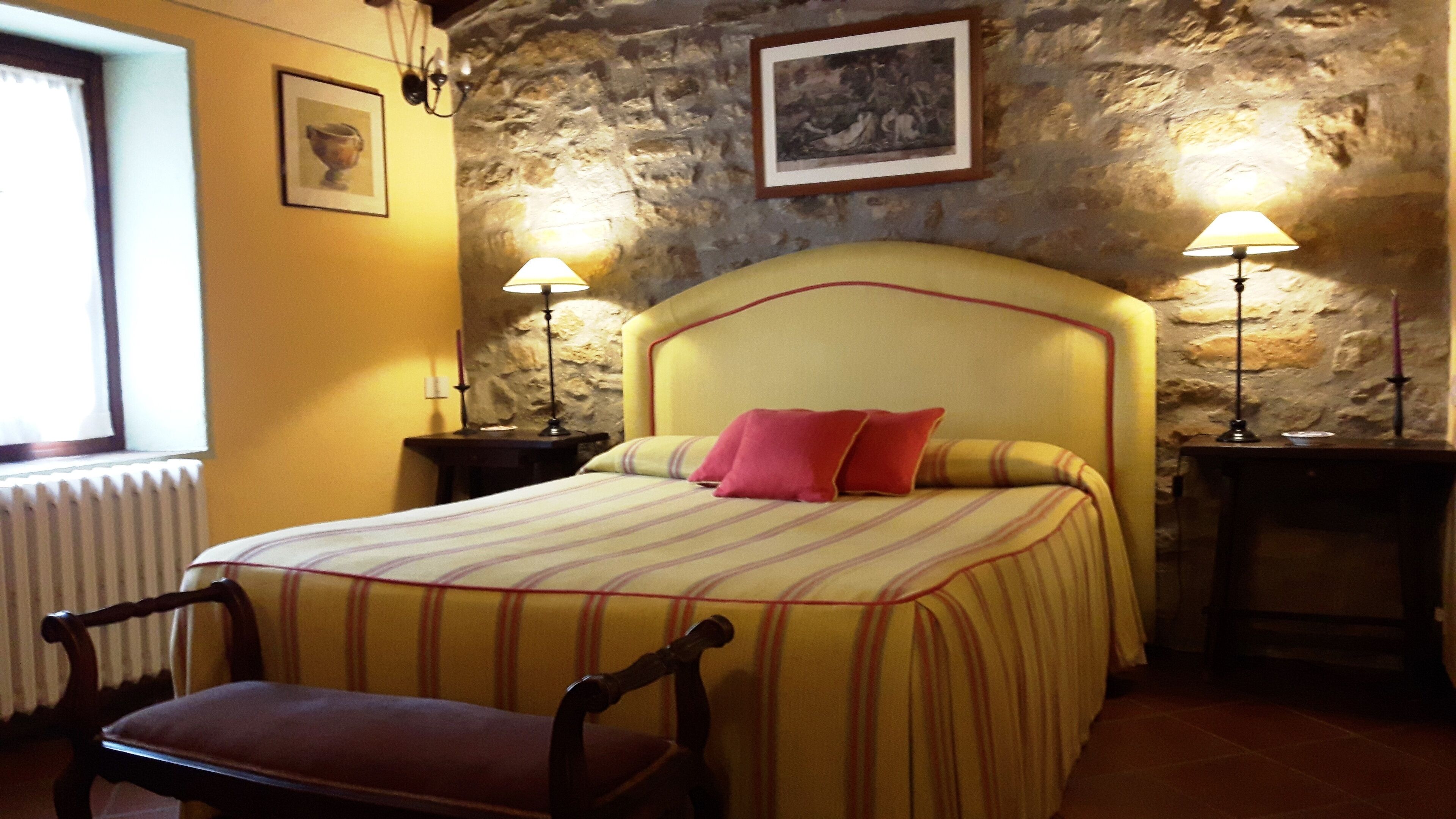 Een bed met een gestreepte dekbedovertrek, een rood kussen en twee bedlampjes. Voor het bed staat een stoel en er is een stenen muur achter.