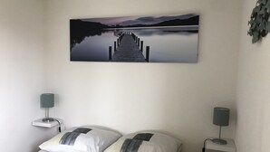 Ein Schlafzimmer mit einem Bett, zwei Nachttischen, einer Wandlampe und einem Wandbild von einem Steg, sowie einer an der Wand befestigten Pflanze.
