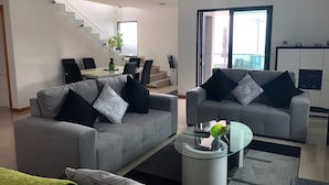 Ein modernes Wohnzimmer mit zwei grauen Sofas, einem Glastisch und einem Essbereich mit Tisch und Stühlen.