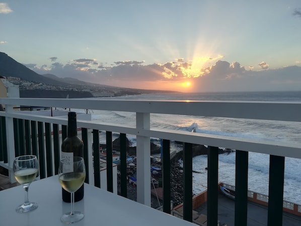 A balcony with a table, two wine glasses, and a bottle of wine. The view includes a sunset over the ocean and a distant town.