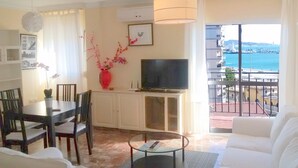 Ein Wohnzimmer mit Fernseher, Esstisch, Stühlen, einer Couch, einem Couchtisch, einer Vase mit Blumen und einem Balkon mit Meerblick.