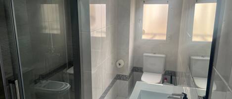 Un bany amb una cabina de dutxa de vidre, un lavabo blanc amb mirall i un vàter amb una finestra a sobre.