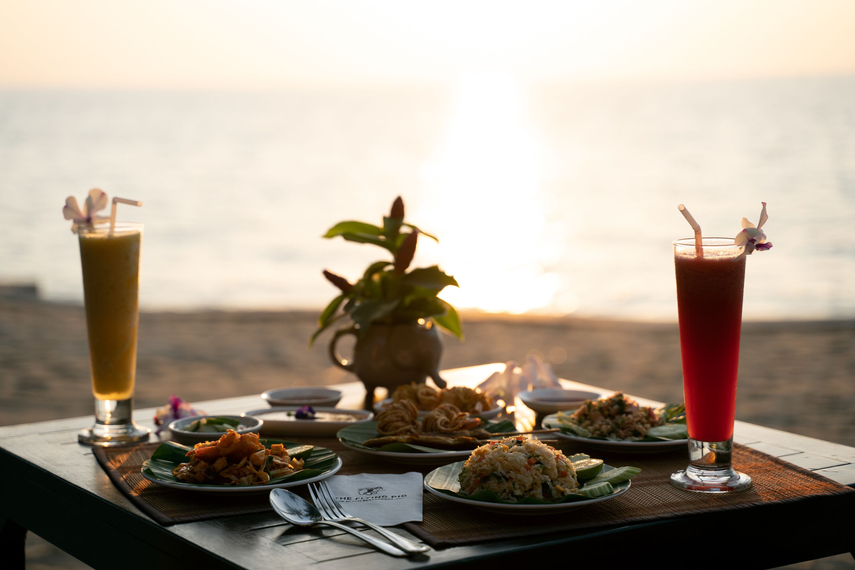 A table set with various dishes and drinks at sunset by the sea.