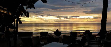 An outdoor seating area with tables and chairs facing a sunset over the ocean.