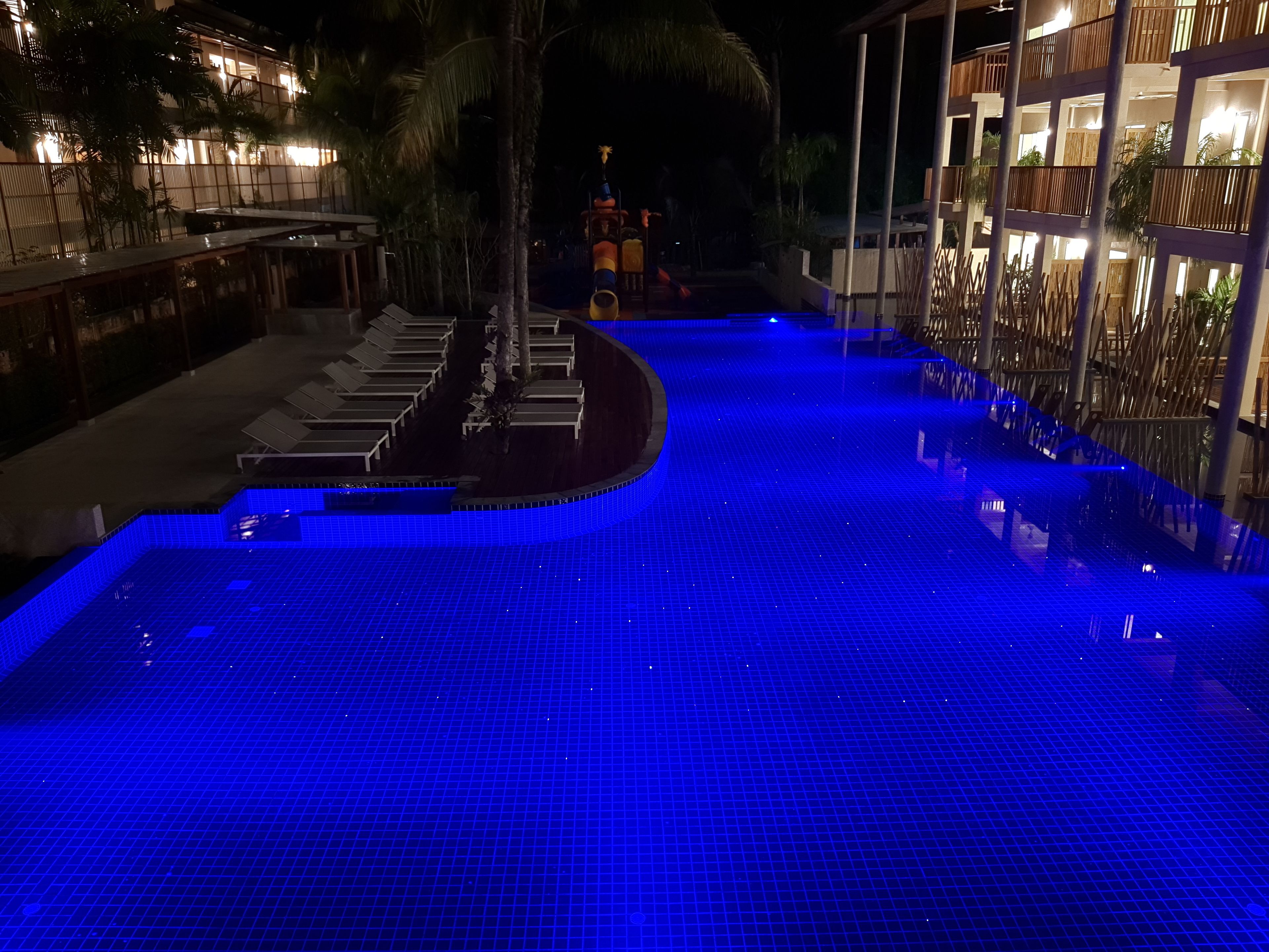 A nighttime swimming pool with blue lighting, surrounded by lounge chairs and a building with balconies.