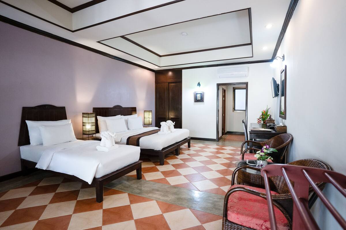 Une chambre d’hôtel avec deux lits, un sol à carreaux, un coin salon avec une chaise et une table, et une porte donnant sur une autre pièce.