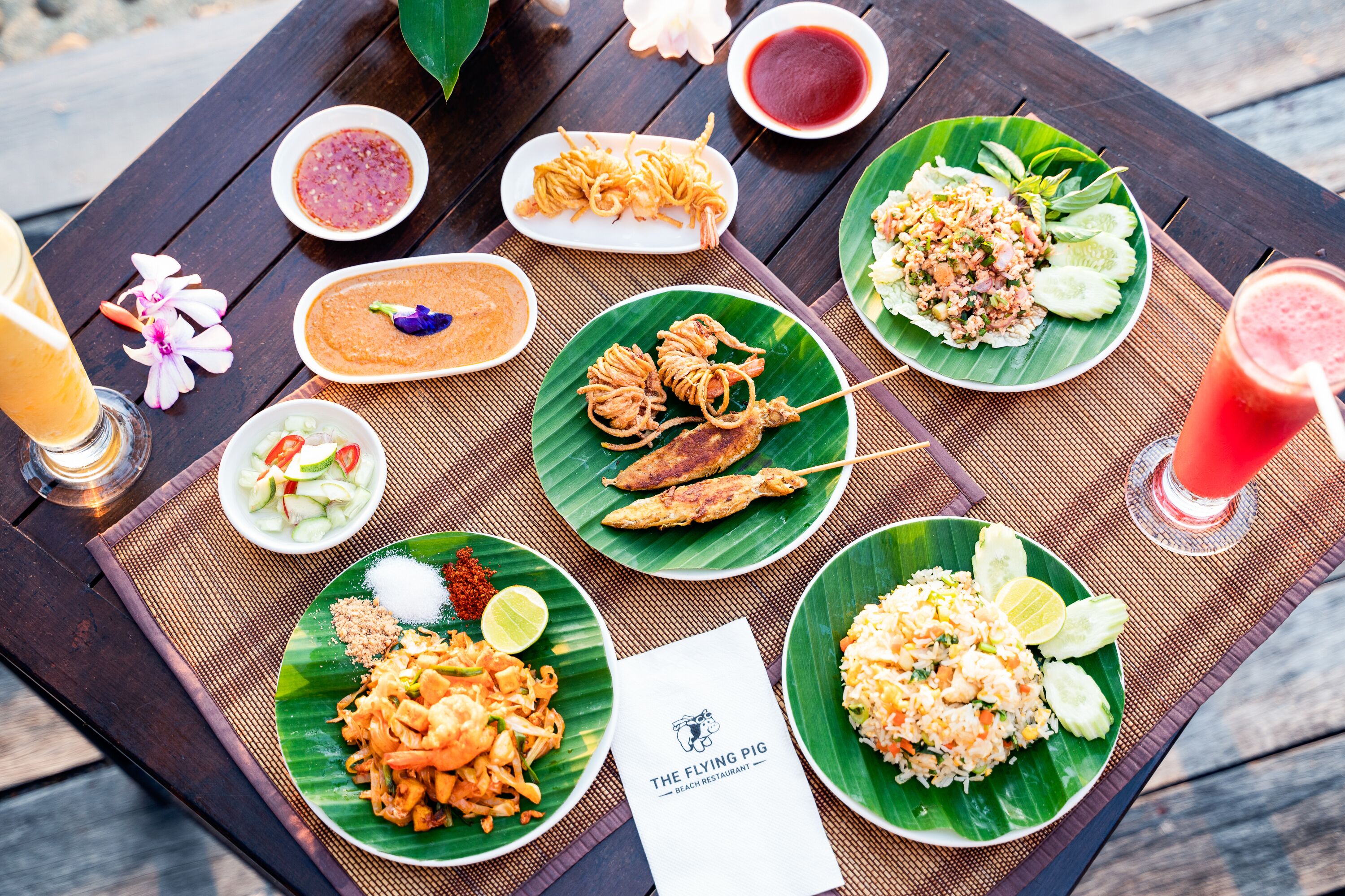 A table with various dishes and drinks, including noodles, fish, and rice, on a wooden table with a placemat.