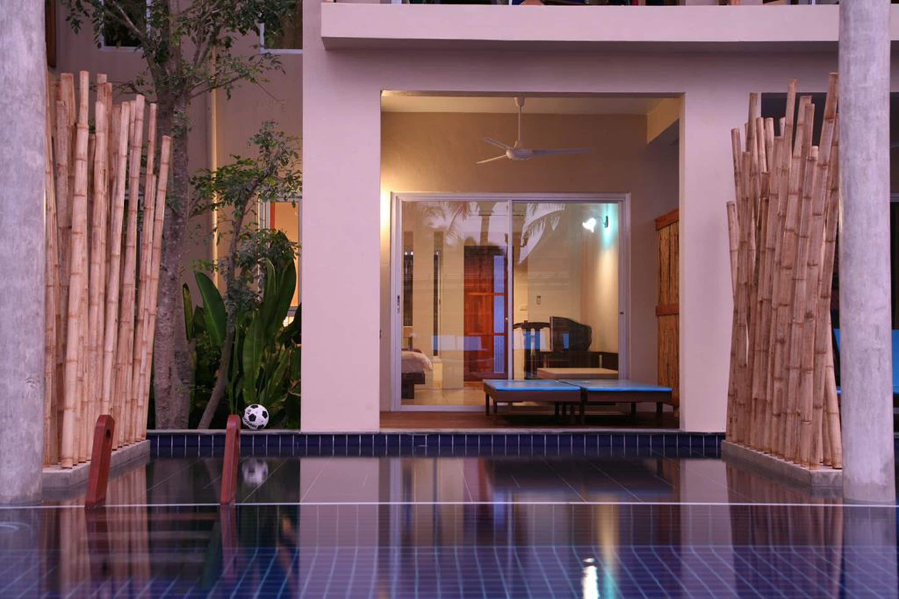 A modern hotel room with a glass door leading to a balcony, a ceiling fan, and a pool area with a bamboo fence.