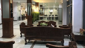 Un hall d’entrée doté d’un espace de repos central, comprenant un banc en bois et des chaises, le tout entouré de murs en verre et agrémenté d’un grand ventilateur.