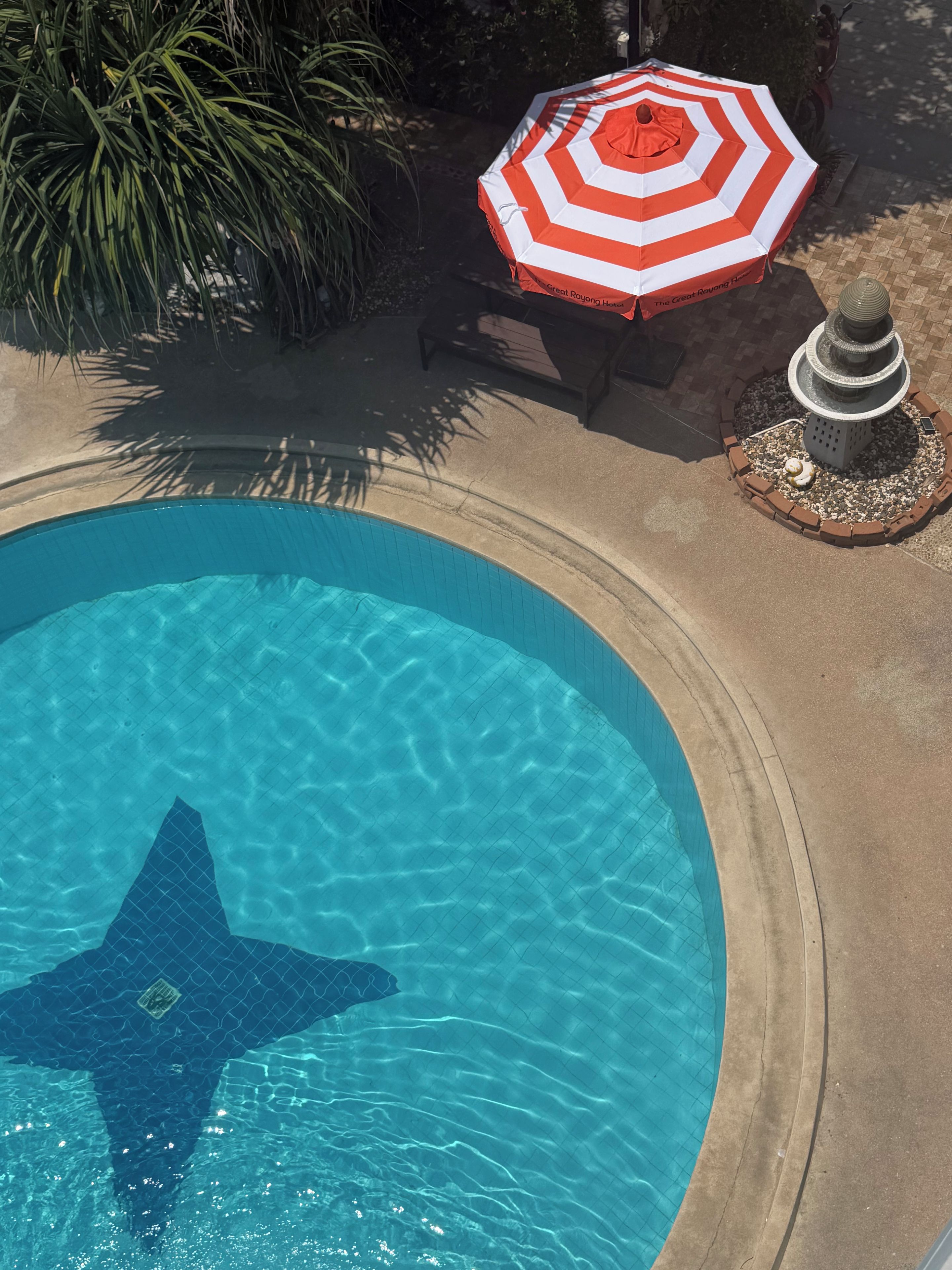 Una piscina con diseño en forma de estrella, rodeada por una solana de hormigón, una sombrilla a rayas rojas y blancas y una fuente decorativa.