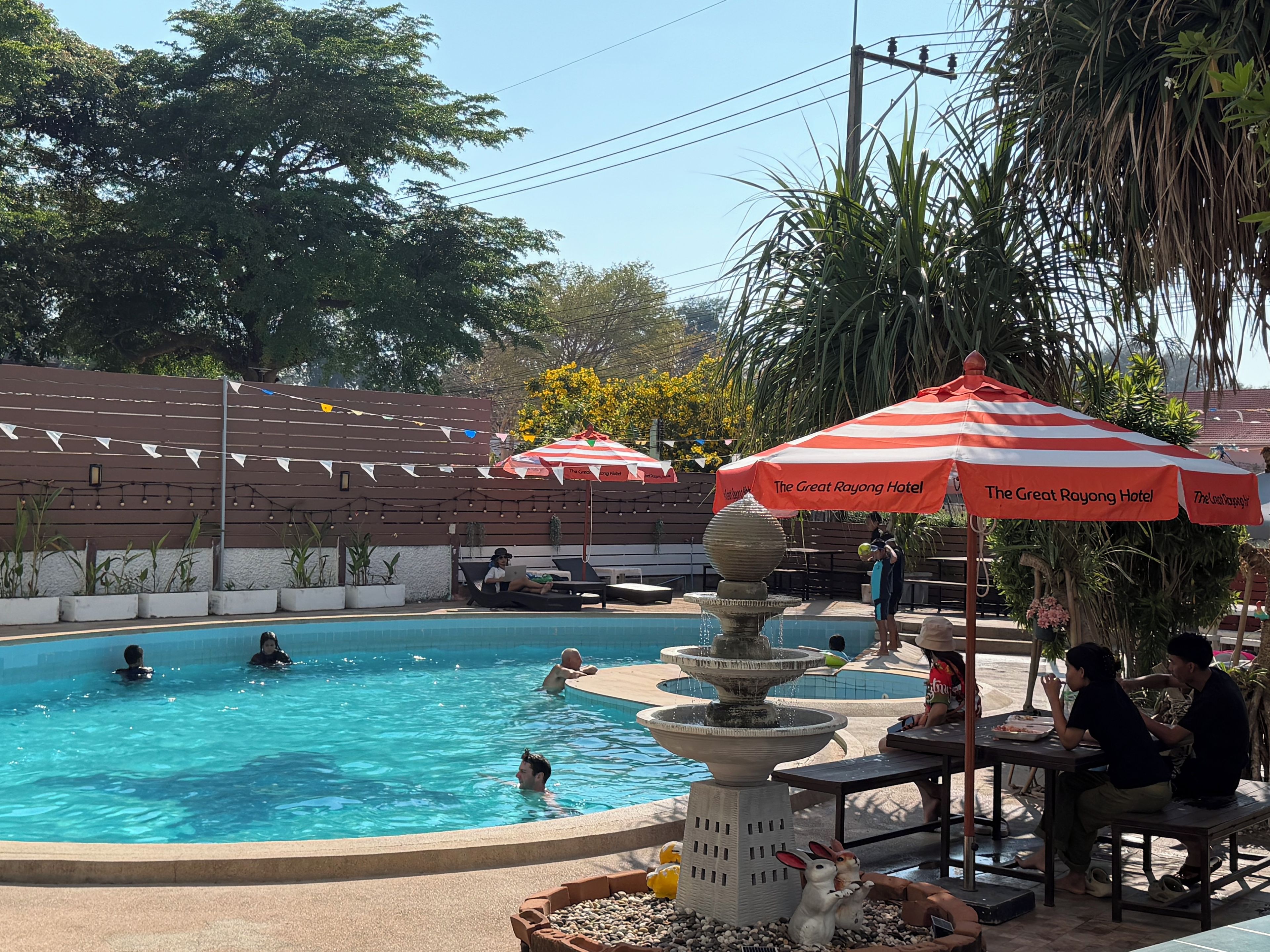 A swimming pool with a fountain, surrounded by lounge chairs and umbrellas, with a brick wall and trees in the background.