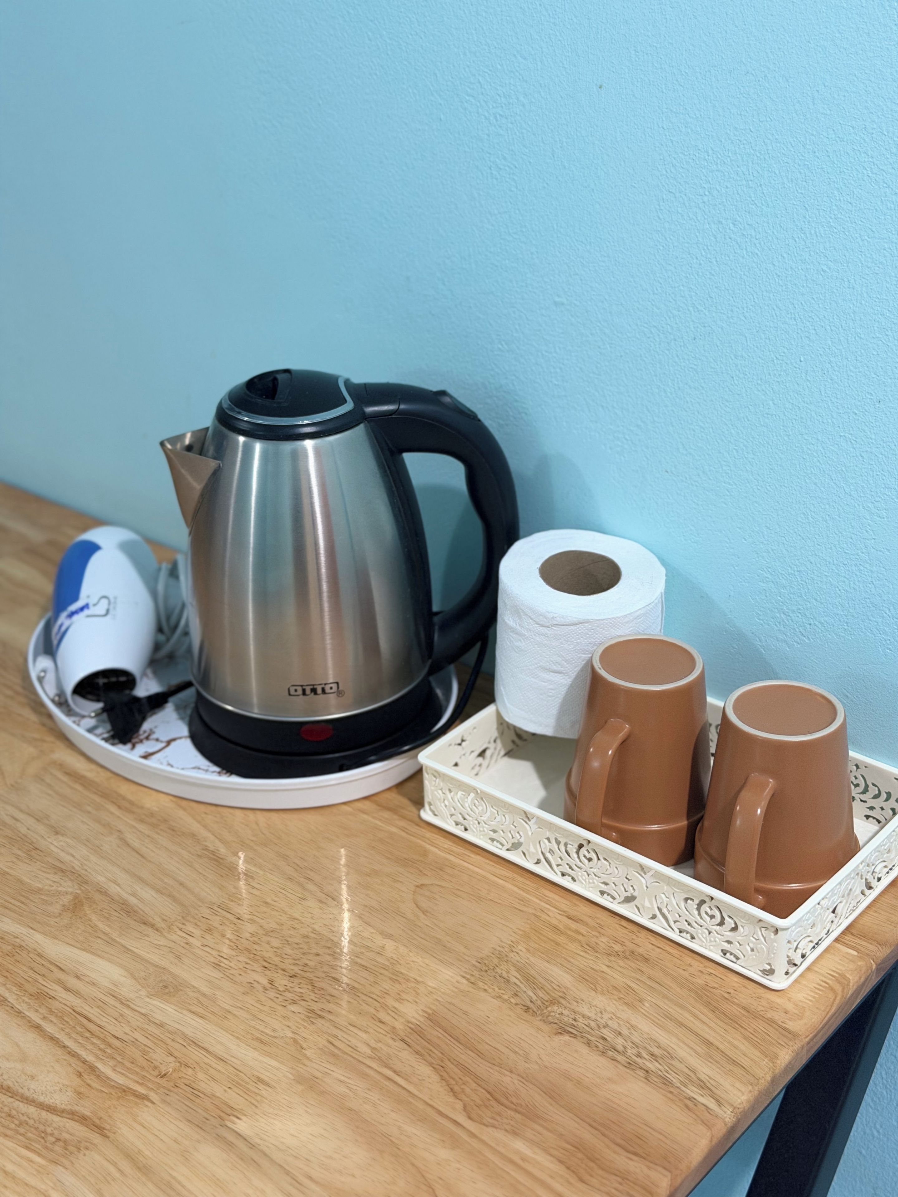 A kettle, two mugs, a roll of toilet paper, and a power adapter on a wooden table against a blue wall.
