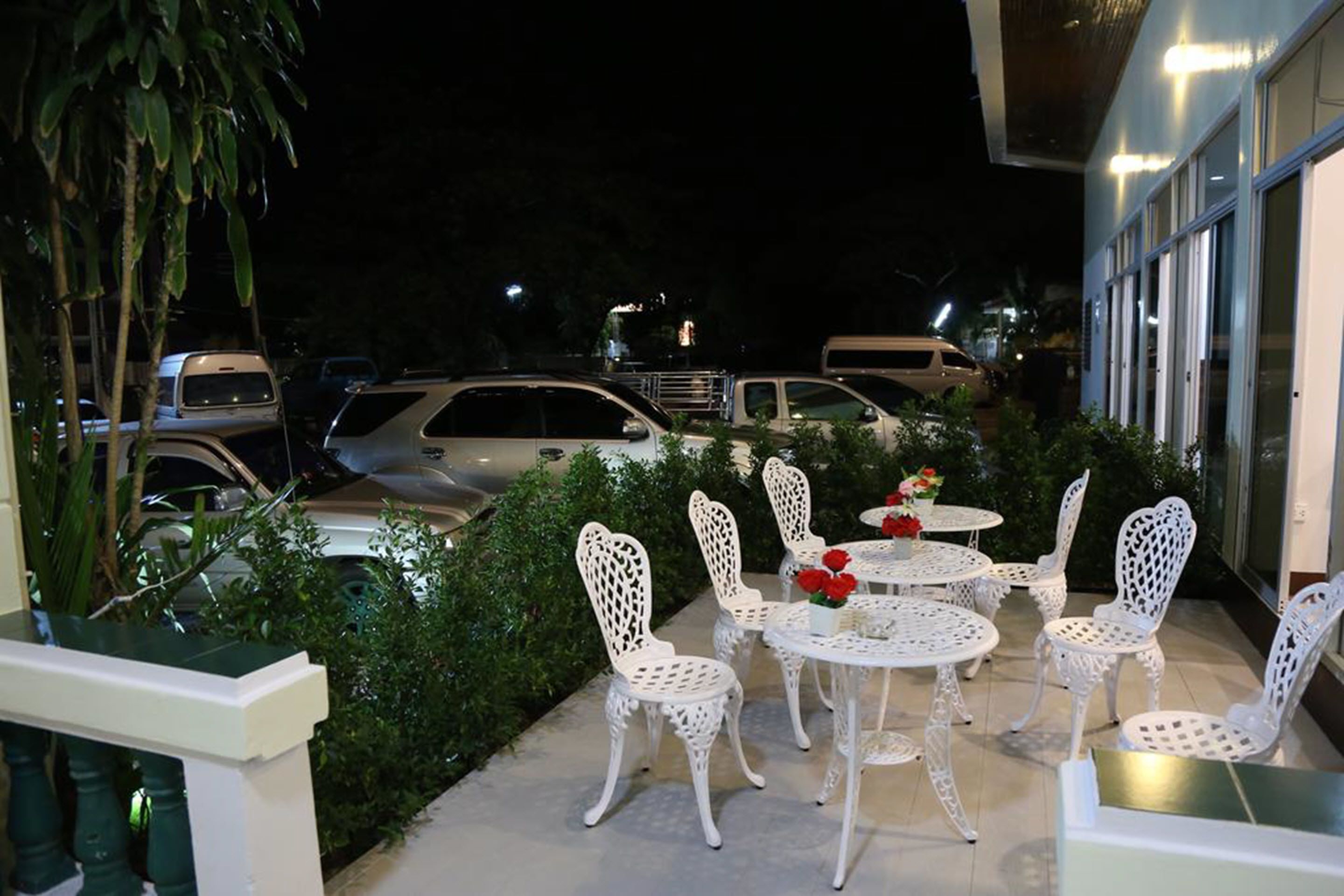 Zona exterior con mesas y sillas de metal blanco, rodeada de vegetación y coches estacionados.