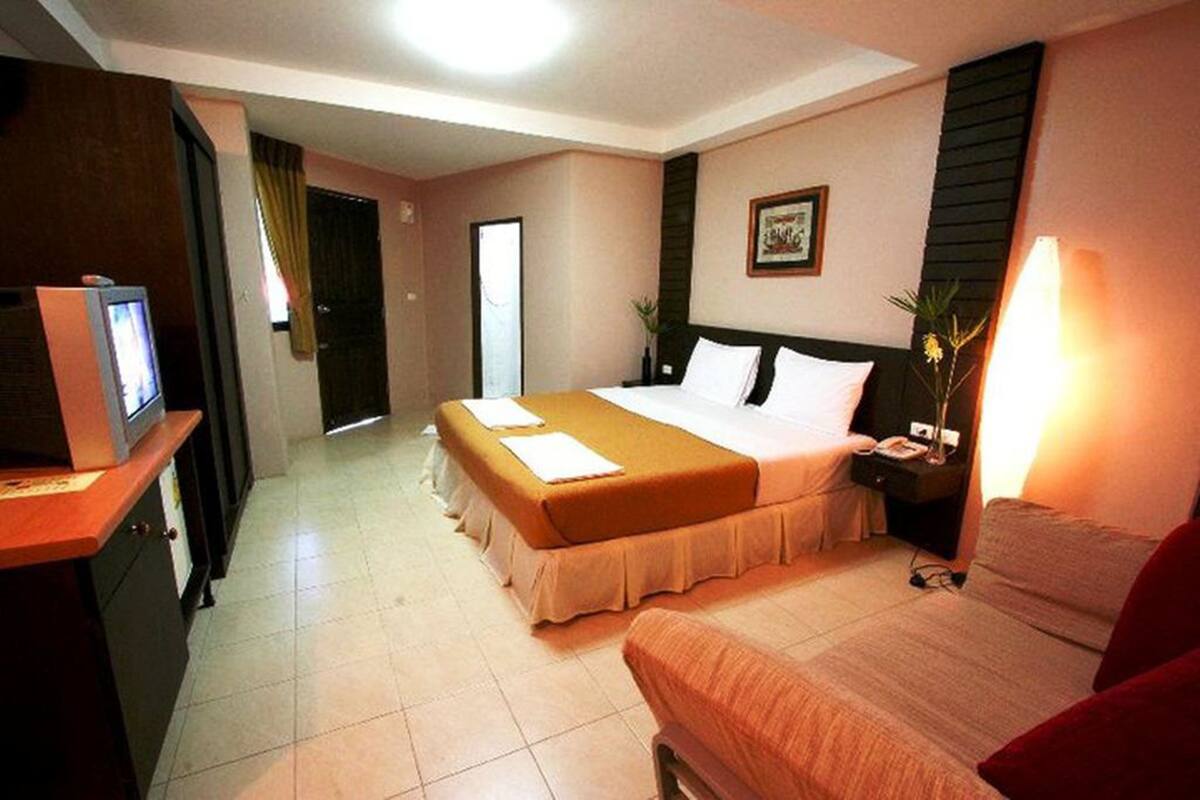 A hotel room with a bed, bedside table, lamp, TV, and a small desk with a computer.