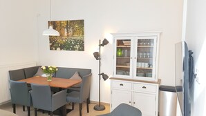 Ruokailutilassa on puinen pöytä, harmaat tuolit, maljakko täynnä kukkia, lattiavalaisin, lasiovilla varustettu kaappi ja seinällä oleva taulu.