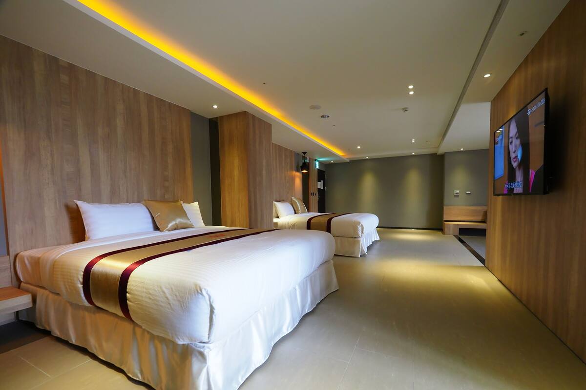 A modern hotel room with a large bed, a smaller bed, a sofa, and a TV on the wall.
