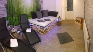 Ein Schlafzimmer mit Steinwand, einem Bett mit Kissen, einem Stuhl, einem kleinen Tisch mit Flasche und Gläsern sowie einem Nachttisch mit Laterne.