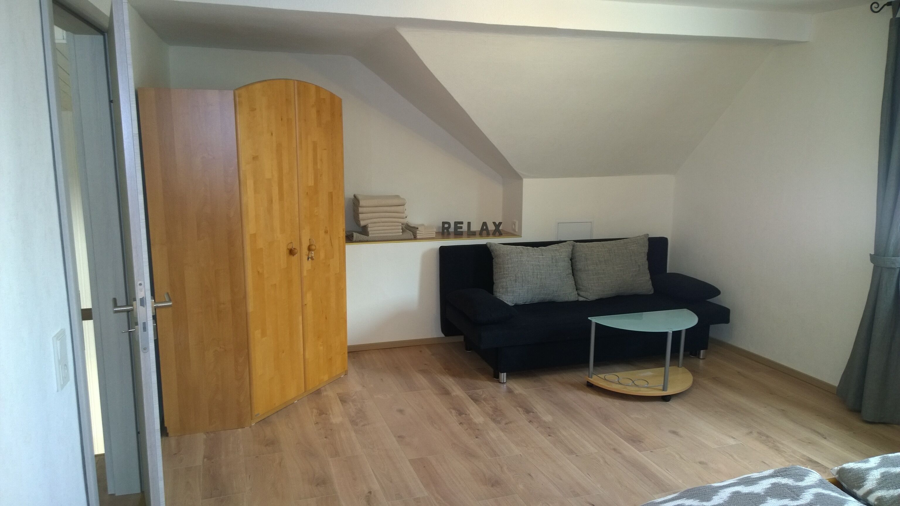 A room with a wooden wardrobe, a black sofa with cushions, a small round table, and a shelf with folded towels and a 'RELAX' sign.