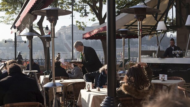 Een terras met tafels en stoelen, parasols en uitzicht op een rivier en gebouwen op de achtergrond.
