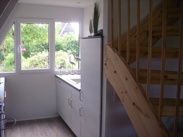 A kitchen with a wooden staircase, a window with a view of greenery, and a refrigerator. - Vacation home close to the beach with patio cover; fiberglass; garden; parking at the house (Damp)