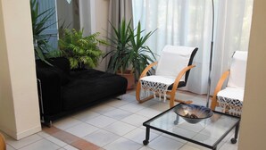 Ein Wohnzimmer mit schwarzem Sofa, zwei weißen Sesseln, einem Glastisch und Topfpflanzen.