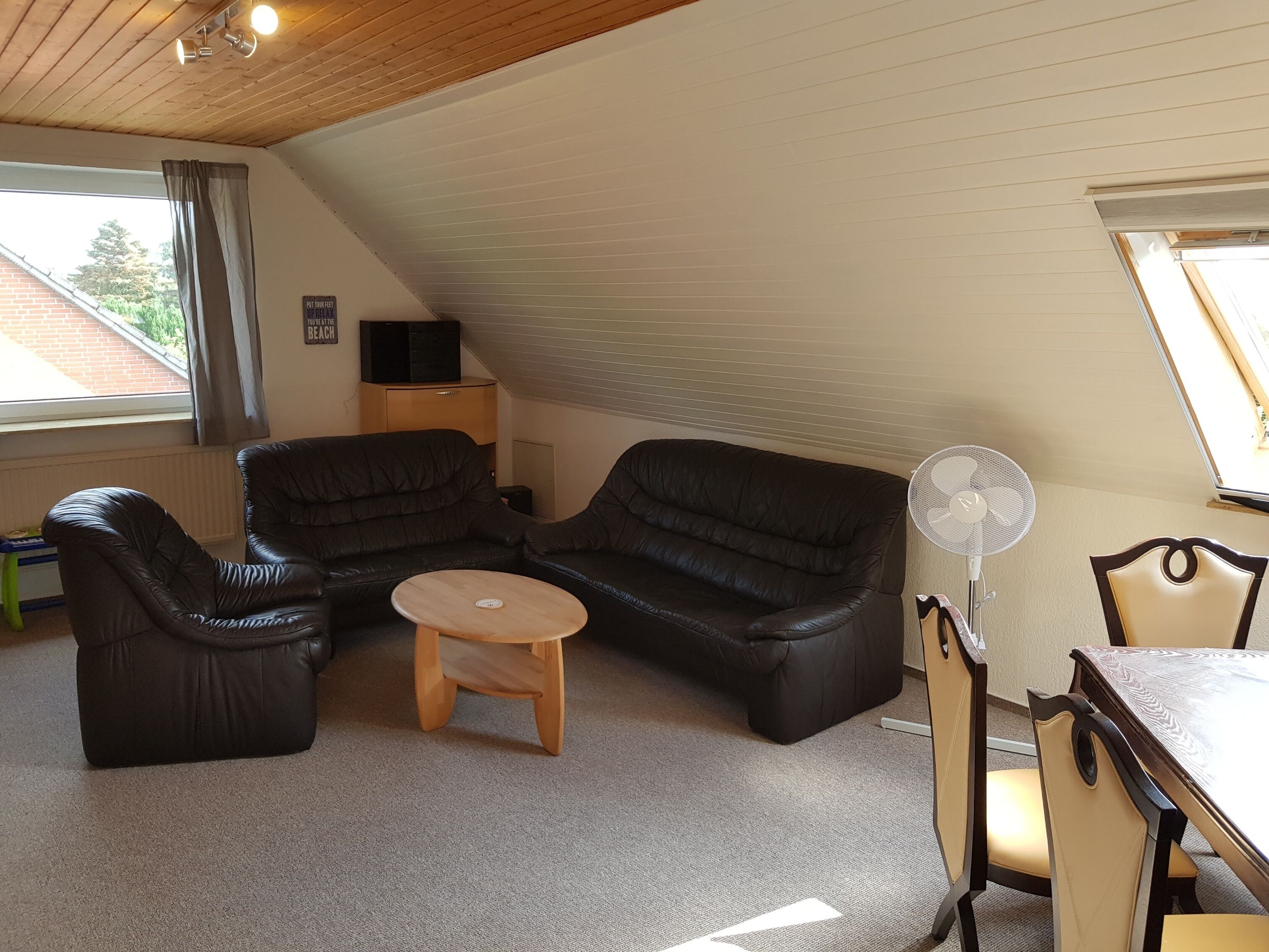 A cozy room with a sloped ceiling, a black leather sofa, a small wooden table, a fan, and a dining table with chairs.