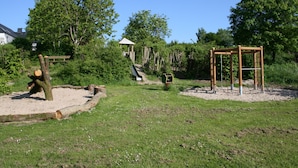 A playground with climbing structures and a slide, surrounded by trees and a house in the background.